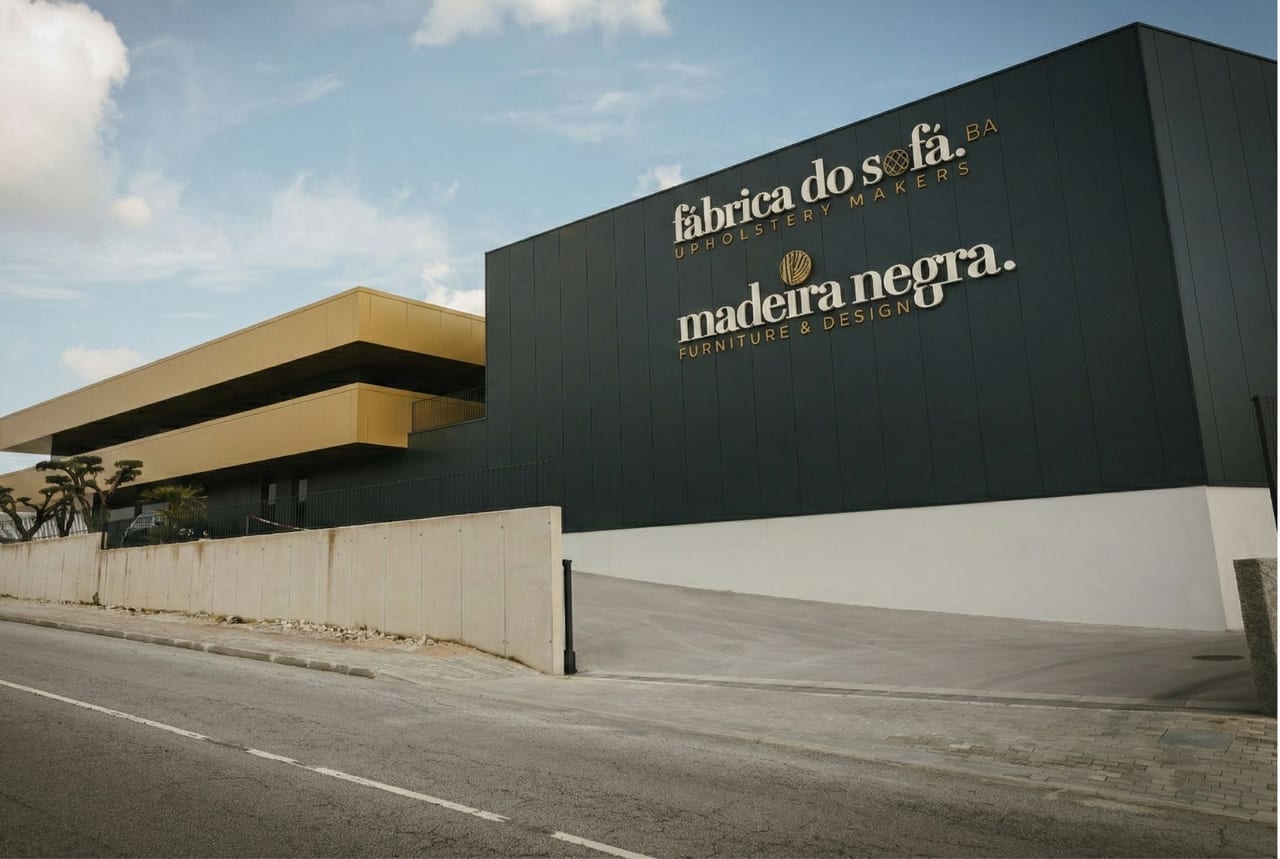 Fachada da fábrica em chapa preta com letras douradas e pala amarela, vista da estrada sob céu azul